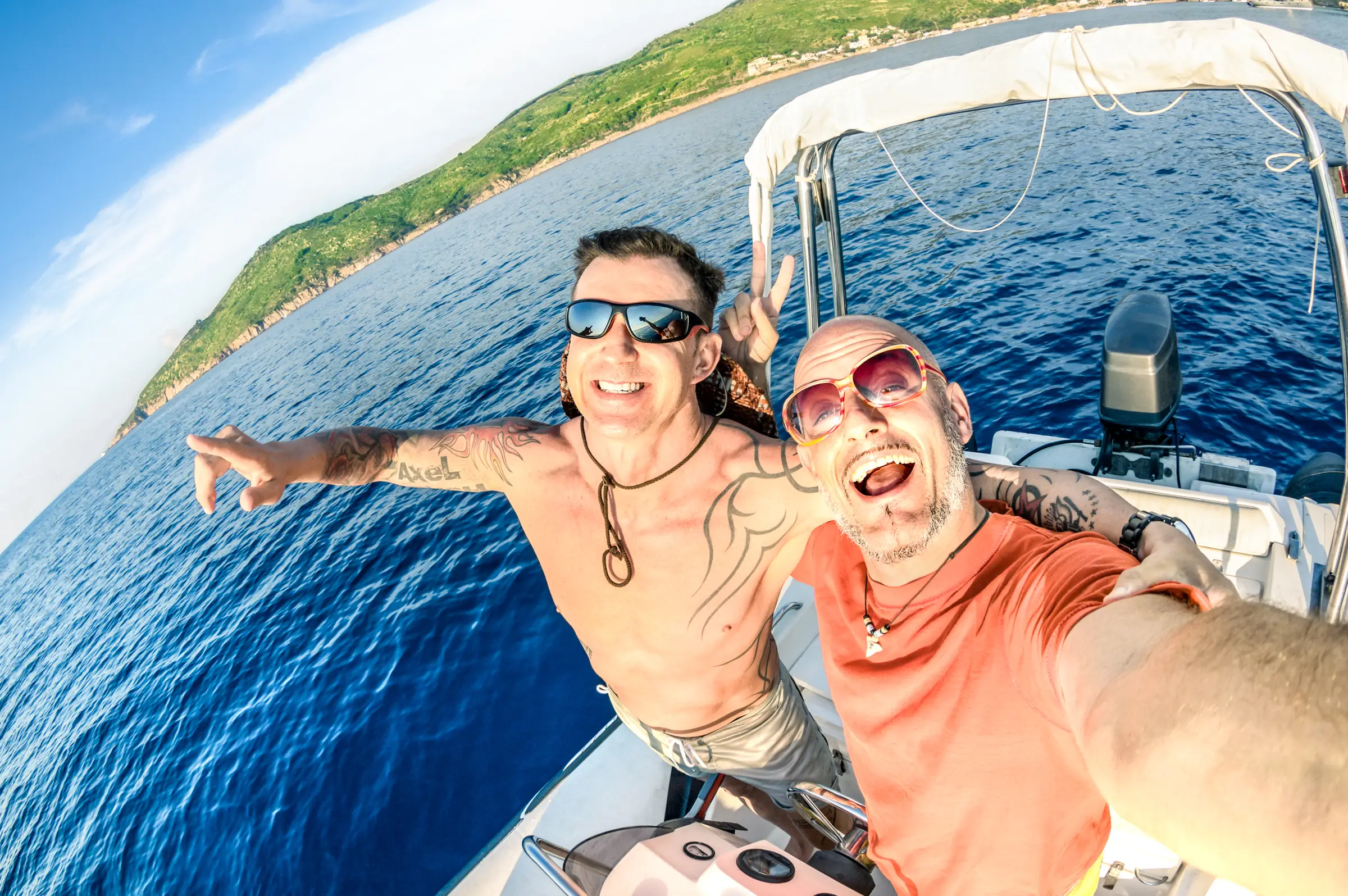 Two grinning men taking a selfie on a speedboat at sea, with a green headland behind
