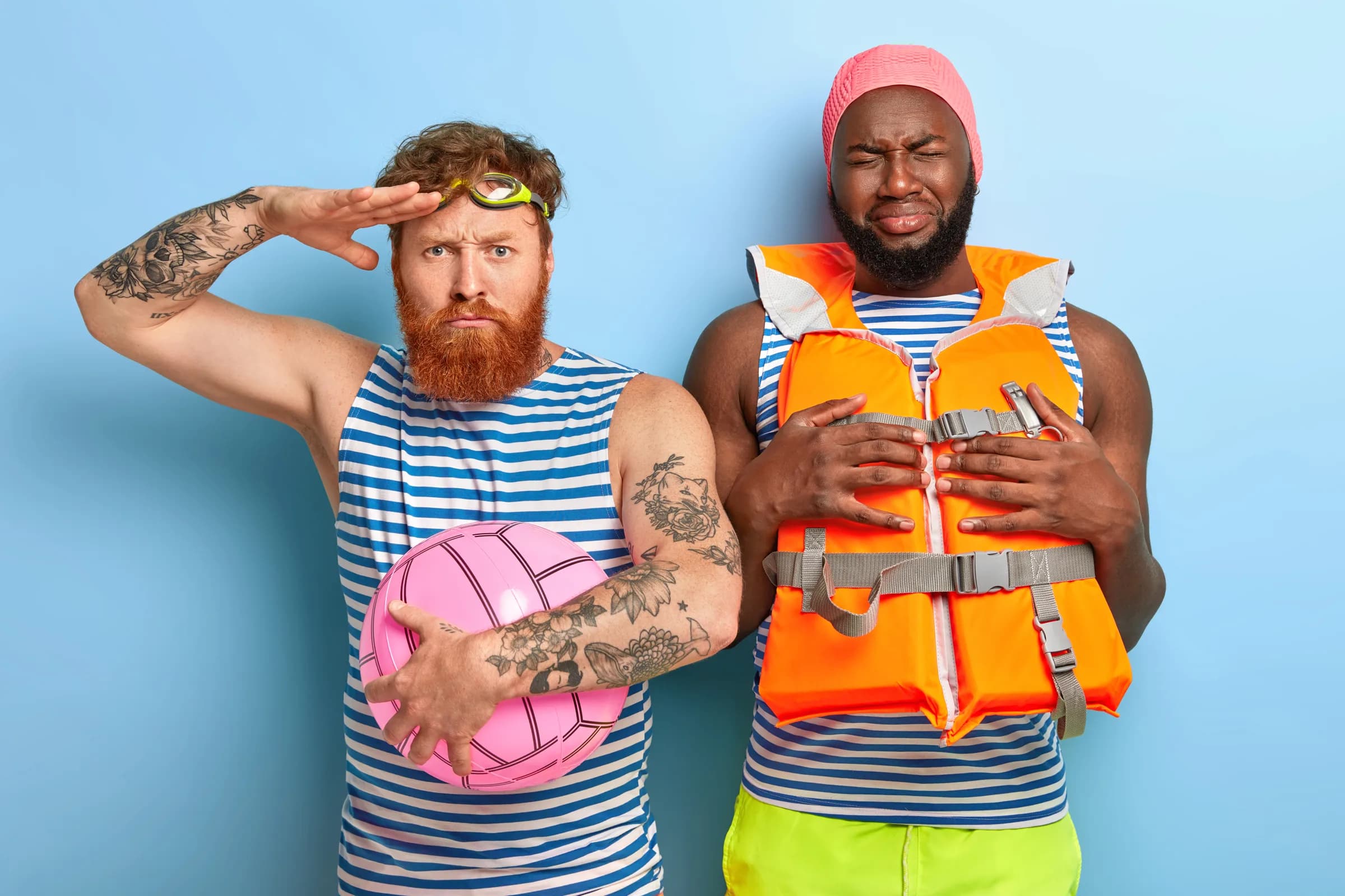 Two bearded men in striped vests against a blue wall, one saluting in swimming goggles with a pink ball, the other hugging an orange life jacket