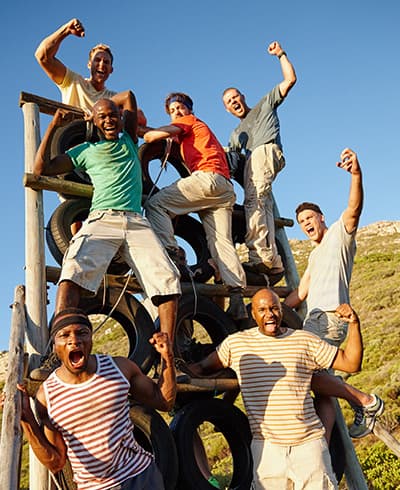 A group of men cheering with fists raised on a wooden and tyre climbing structure outdoors